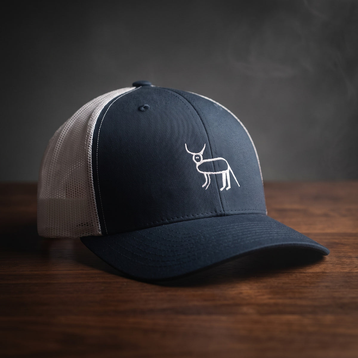 Casquette trucker bleu marine et blanche avec la perruche ratée brodée en blanc, posée sur un bois sombre