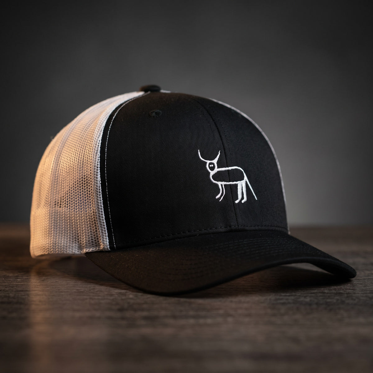 Casquette trucker noire et blanche avec la perruche ratée brodée en blanc, posée sur un bois sombre