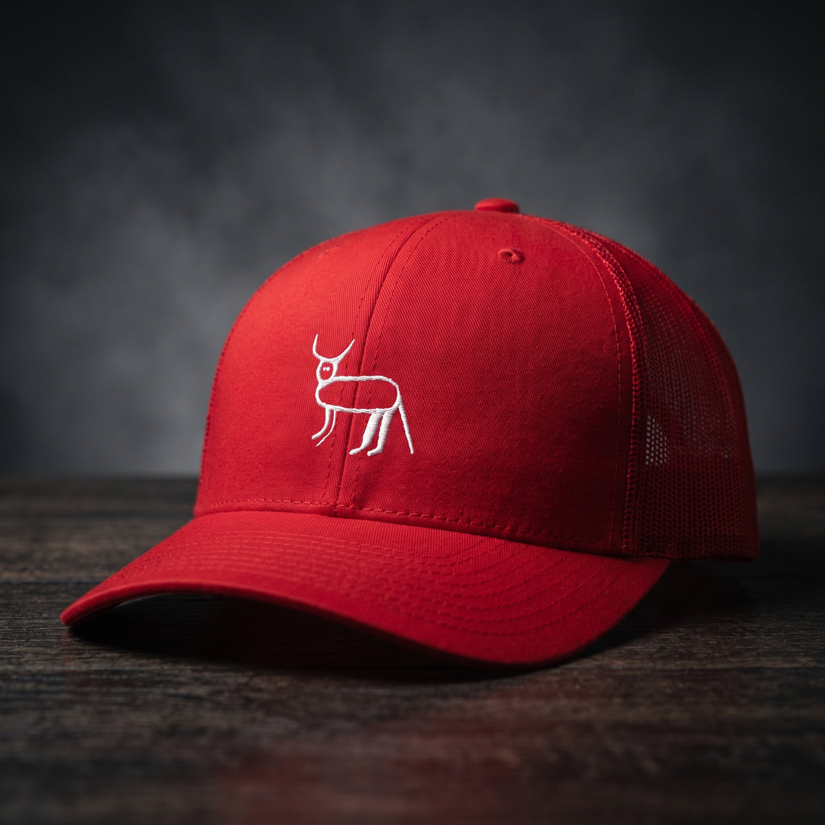 Casquette trucker rouge avec la perruche ratée brodée en blanc, posée sur un bois sombre