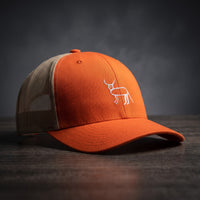 Casquette trucker roux et kaki avec la perruche ratée brodée en blanc, posée sur un bois sombre