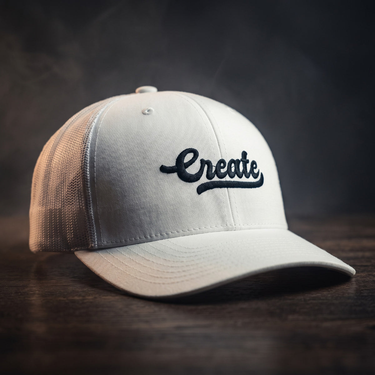 Casquette trucker blanche avec le mot Create brodé en noir en écriture manuscrite rétro, posée sur un plan de bois sombre