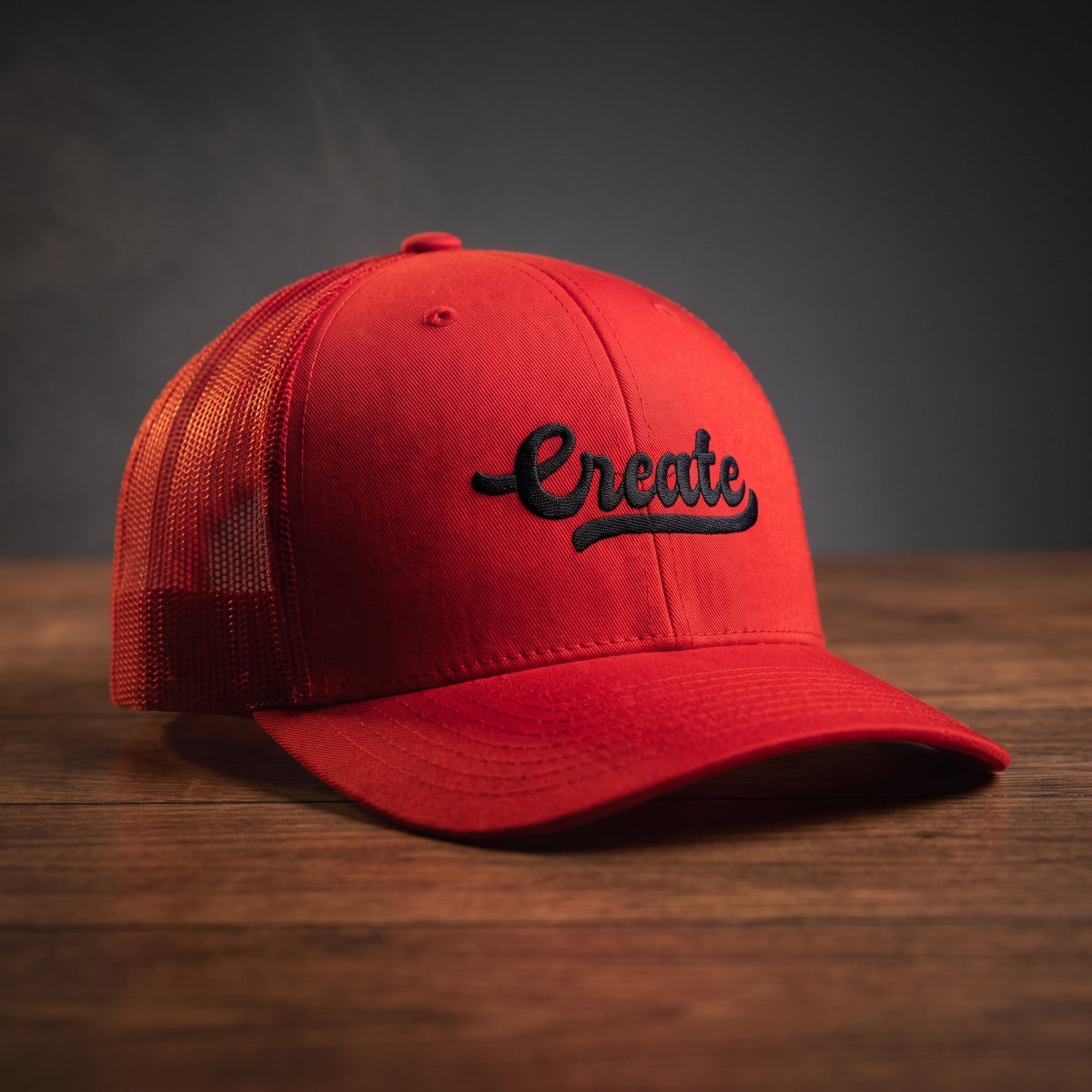 Casquette trucker rouge avec le mot Create brodé en noir en écriture manuscrite rétro, posée sur un plan de bois sombre