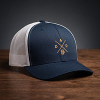 Casquette trucker brodée DAD bleu marine et blanc posée sur bois – logo chope de bière doré