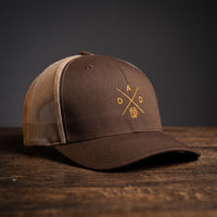 Casquette trucker brodée DAD marron et kaki posée sur bois – logo chope de bière doré