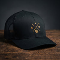 Casquette trucker brodée DAD noire posée sur bois – logo chope de bière doré
