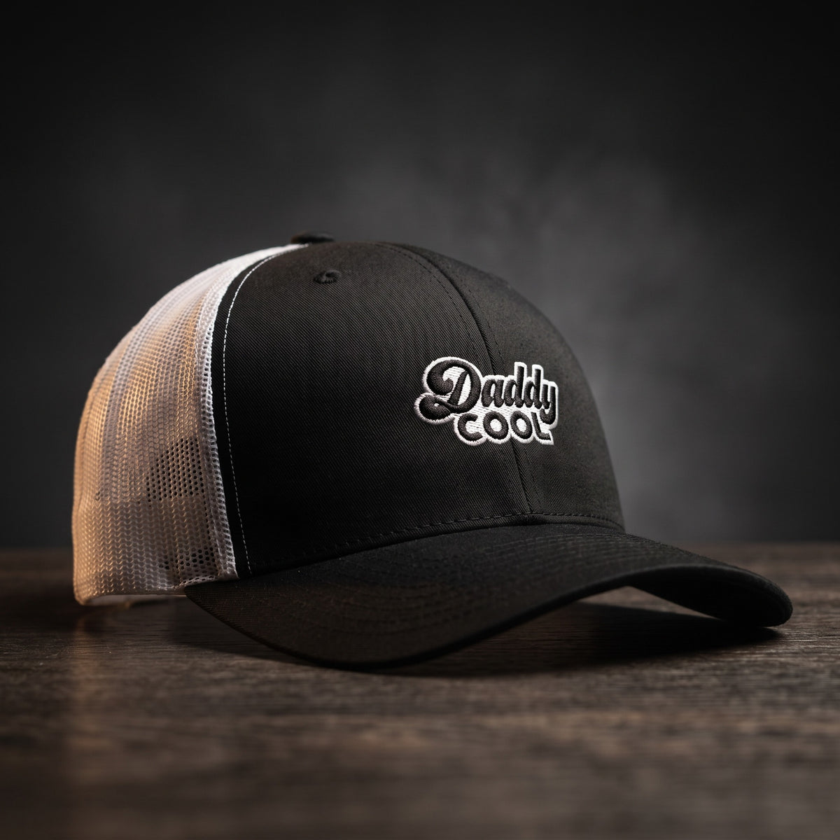 Casquette trucker Daddy Cool noire et blanche, filet blanc, logo brodé, posée sur bois sombre