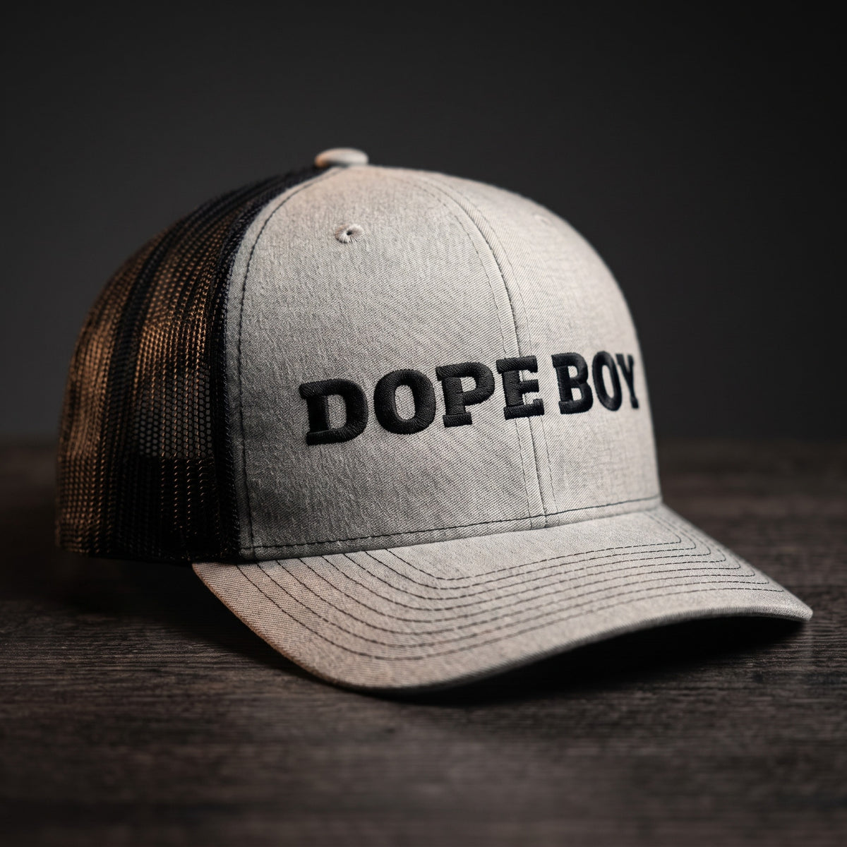 Casquette trucker gris chiné et noire brodée DOPE BOY en lettres noires, posée sur une table en bois sombre