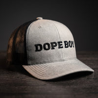 Casquette trucker gris chiné et noire brodée DOPE BOY en lettres noires, posée sur une table en bois sombre