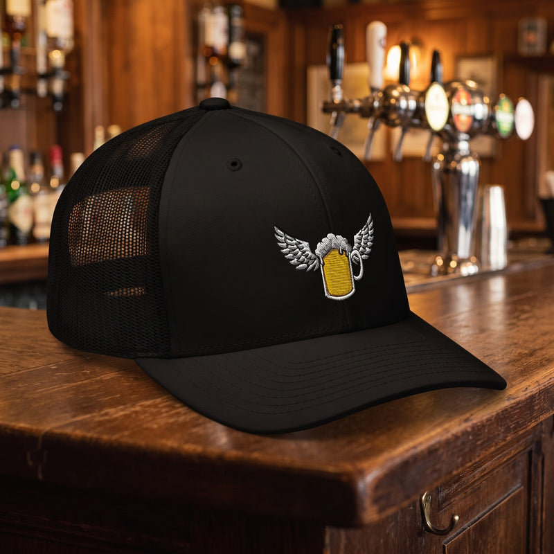 Casquette trucker noire brodée d'une chope de bière dorée avec des ailes blanches posée sur un comptoir en bois d'un pub belge avec des tireuses à bière floues derrière, lumière chaude - CHT'M Boutique