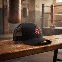 Casquette trucker noire gants de boxe posée sur un banc en bois dans une vieille salle de boxe, sac de frappe et ring en arrière-plan