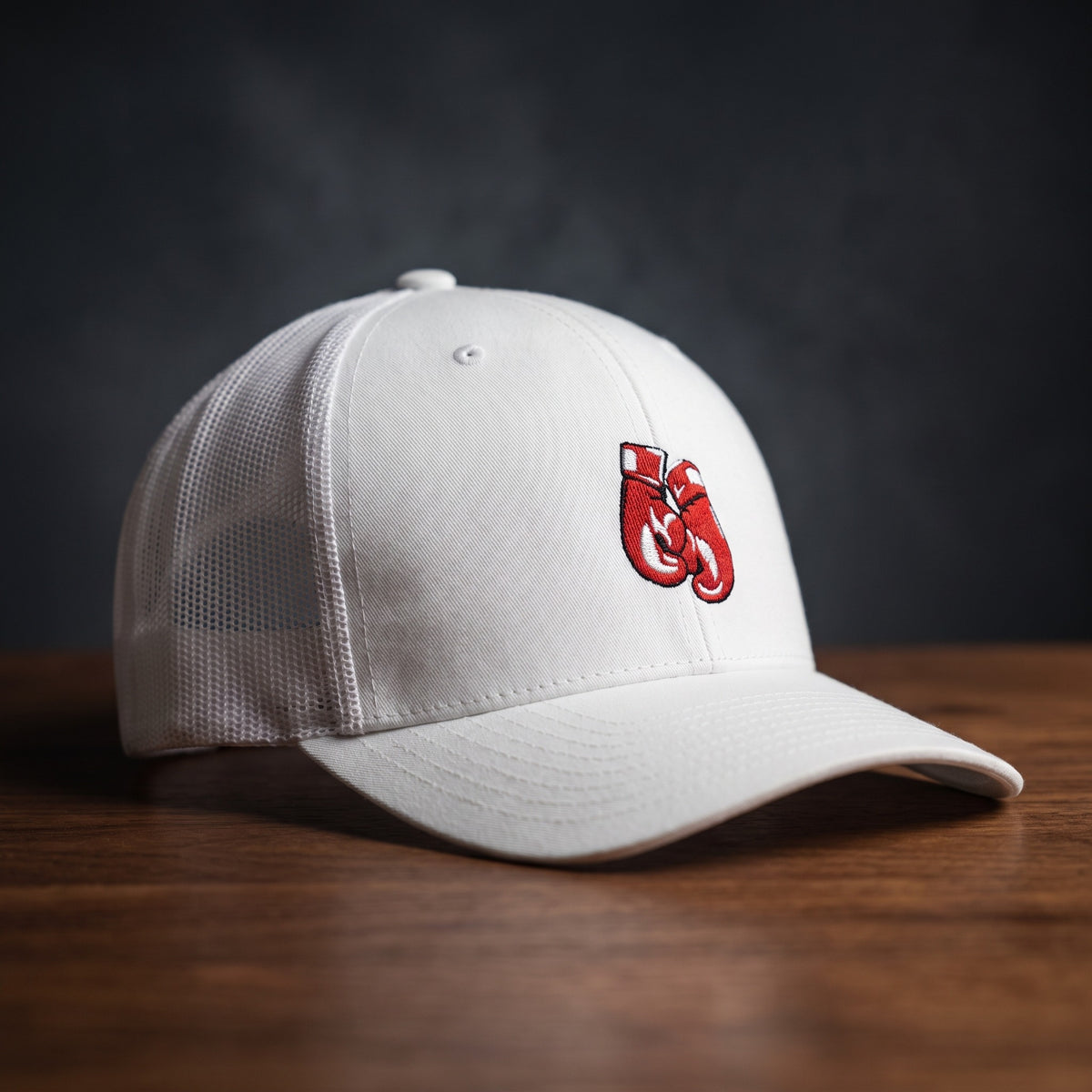 Casquette trucker blanche avec gants de boxe rouges brodés, posée sur un plan de bois sombre