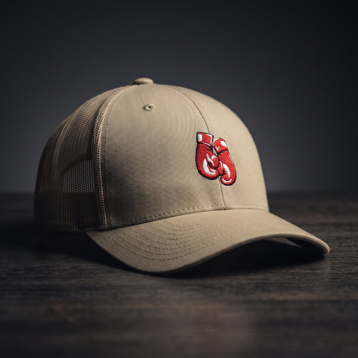 Casquette trucker kaki avec gants de boxe rouges brodés, posée sur un plan de bois sombre