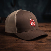 Casquette trucker marron et kaki avec gants de boxe rouges brodés, posée sur un plan de bois sombre