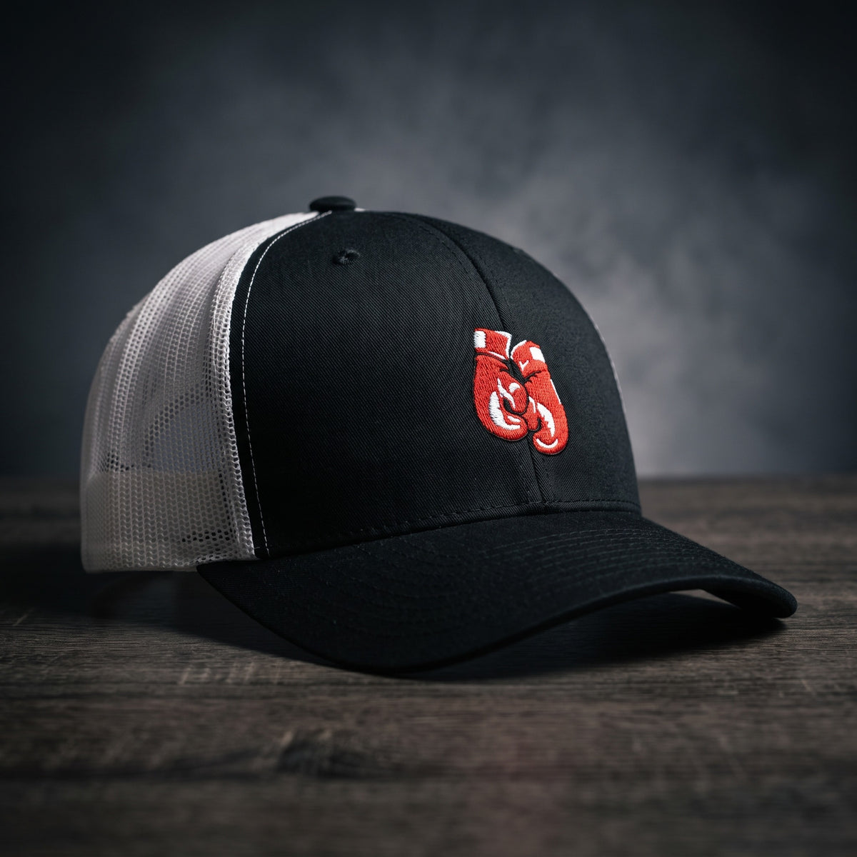 Casquette trucker noire et blanche avec gants de boxe rouges brodés, posée sur un plan de bois sombre