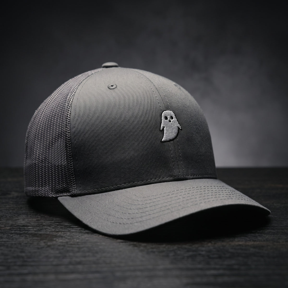 Casquette trucker GHOST gris anthracite avec fantôme blanc brodé, posée sur bois sombre