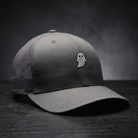 Casquette trucker GHOST gris anthracite avec fantôme blanc brodé, posée sur bois sombre