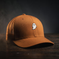 Casquette trucker GHOST caramel avec petit fantôme blanc brodé, posée sur bois sombre