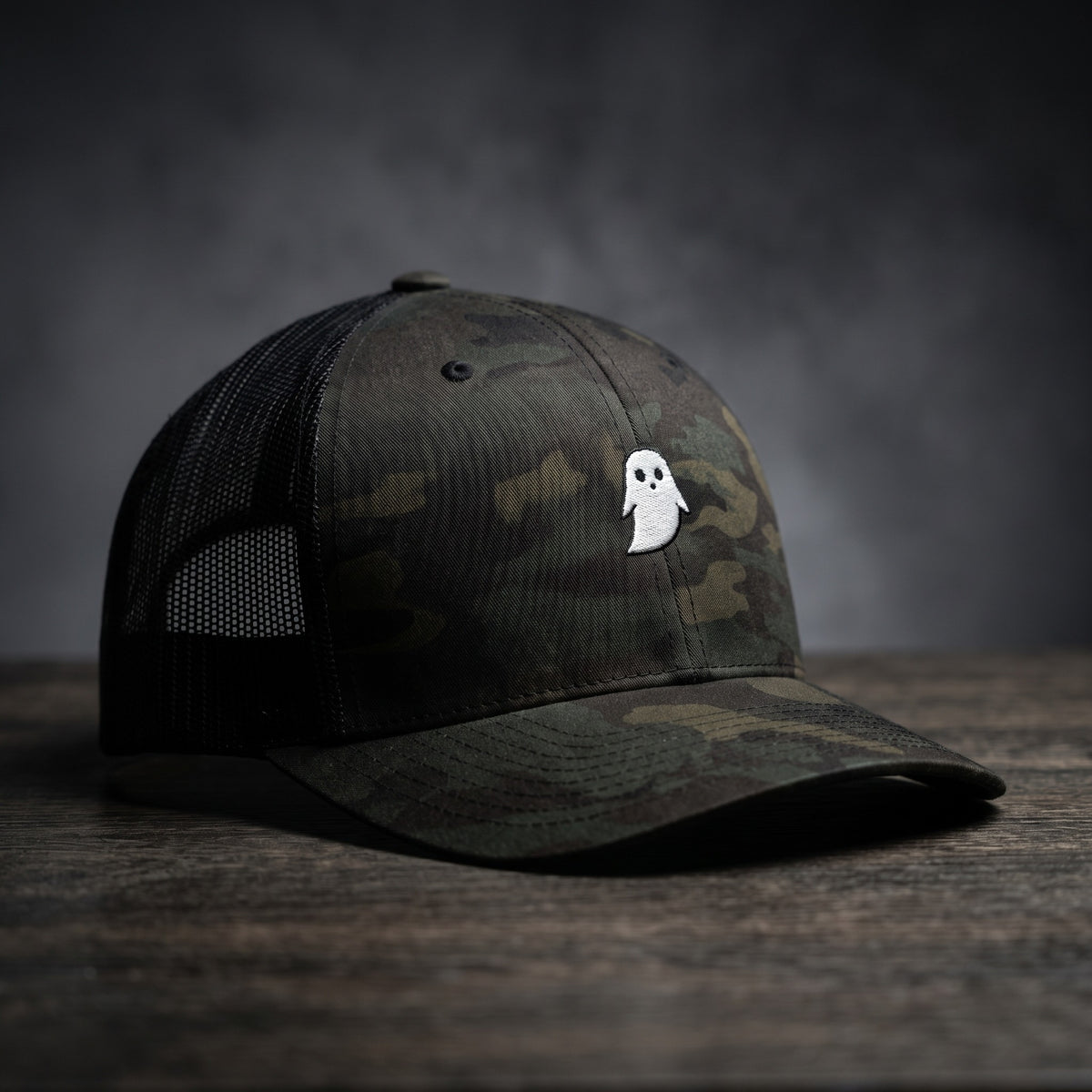 Casquette trucker GHOST camouflage multicam noir avec fantôme blanc brodé, filet noir, sur bois sombre