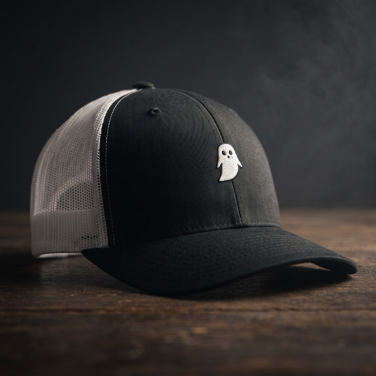 Casquette trucker GHOST noire et blanche avec fantôme brodé, filet blanc, posée sur bois sombre