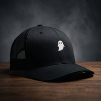 Casquette trucker GHOST noire avec petit fantôme blanc brodé, filet noir, posée sur bois sombre