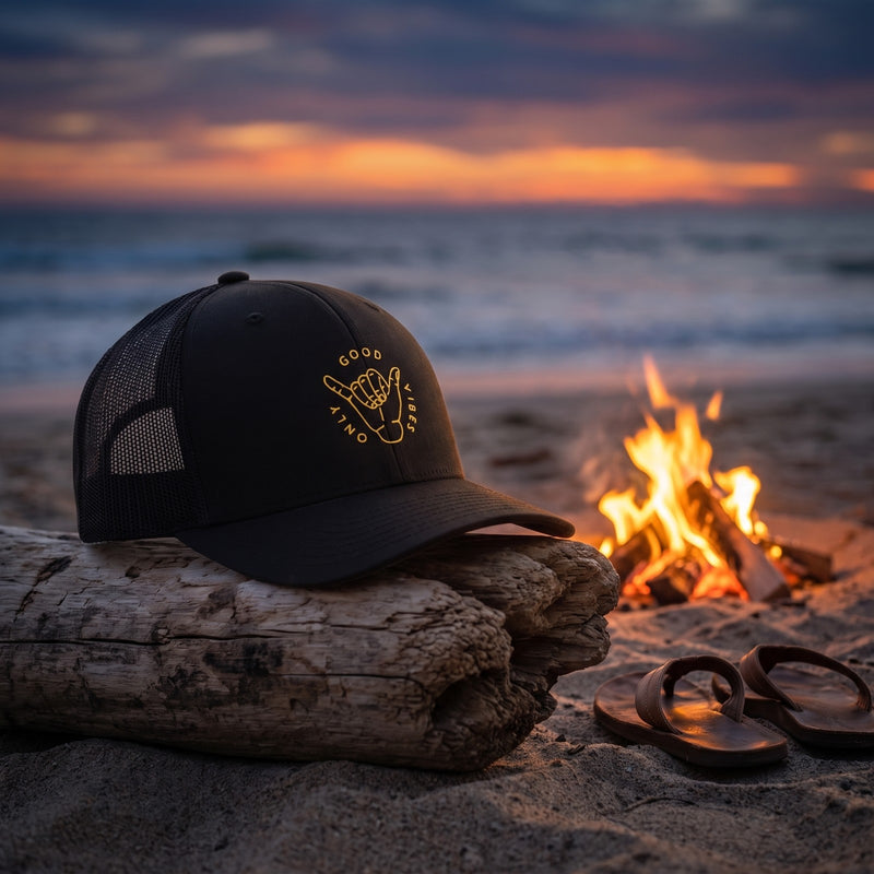 Casquette trucker noire brodée d'un badge rond doré GOOD VIBES ONLY avec une main shaka posée sur un tronc d'arbre flotté sur une plage au crépuscule à côté d'un petit feu et de tongs - CHT'M Boutique