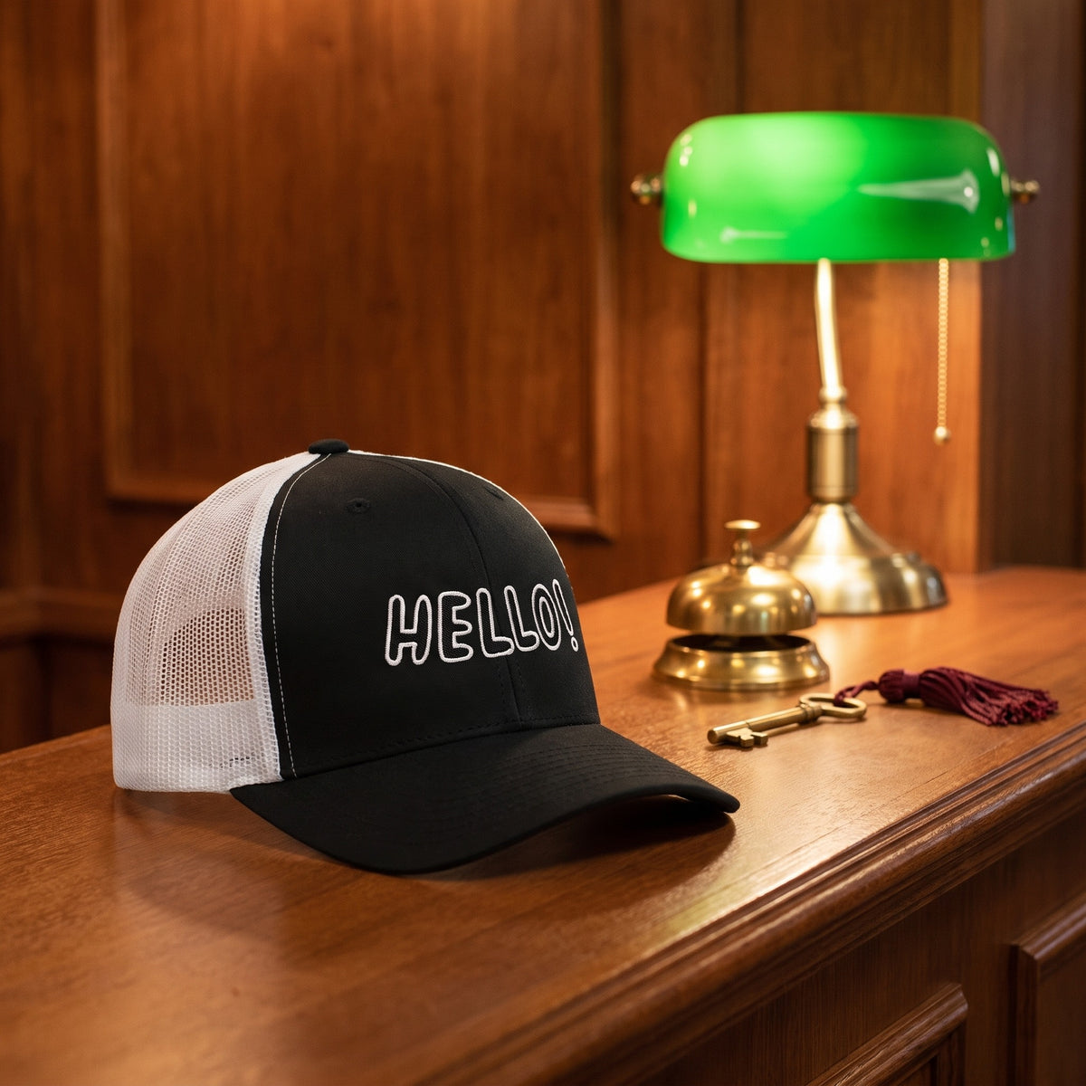 Casquette trucker Hello! posée sur un comptoir de réception d'hôtel avec sonnette en laiton et lampe verte