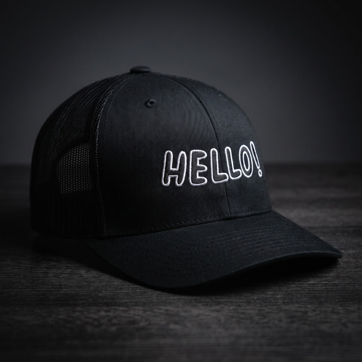 Casquette trucker noire brodée Hello! en lettres blanches, posée sur un plan en bois sombre