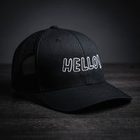 Casquette trucker noire brodée Hello! en lettres blanches, posée sur un plan en bois sombre