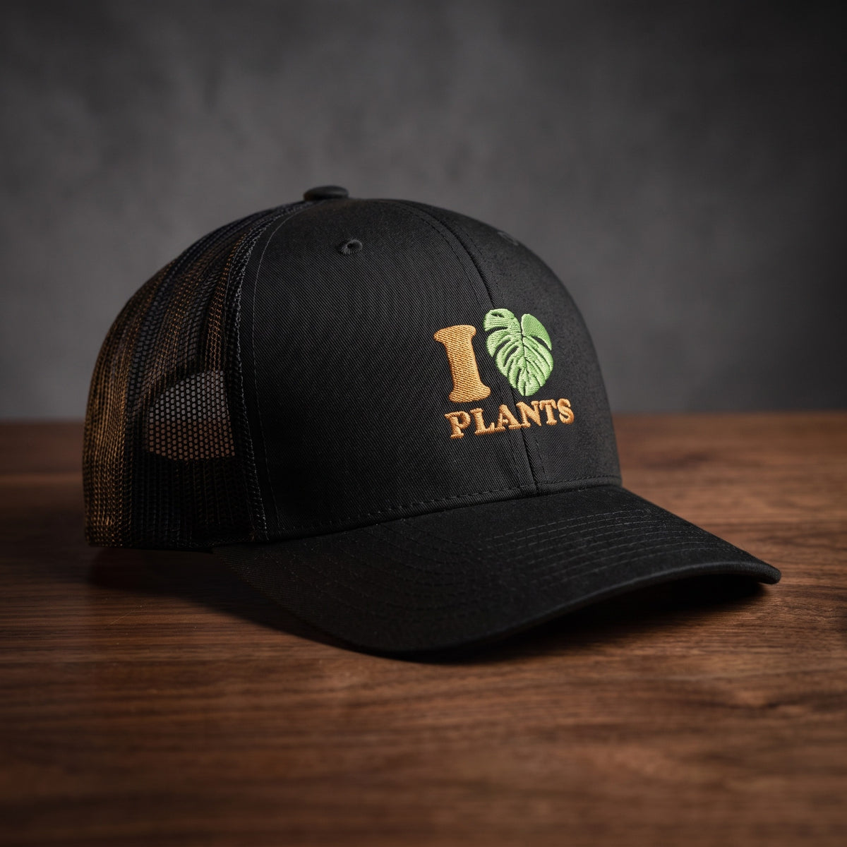 Casquette trucker noire brodée I LOVE PLANTS avec cœur en feuille de monstera verte, posée sur une table en bois sombre