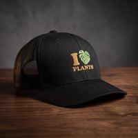 Casquette trucker noire brodée I LOVE PLANTS avec cœur en feuille de monstera verte, posée sur une table en bois sombre