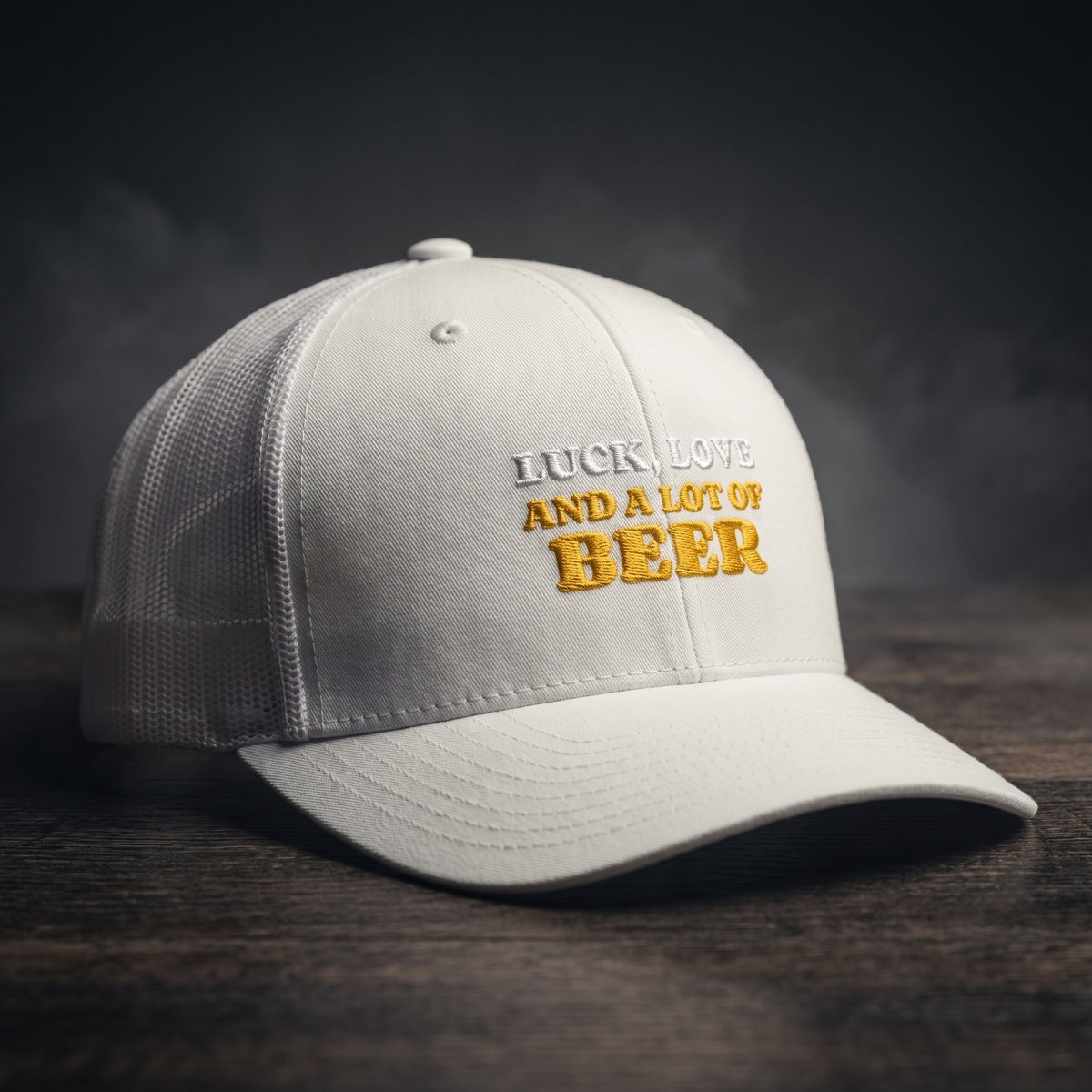 Casquette trucker blanche brodée « Luck, Love and a lot of Beer » en blanc et jaune doré, posée sur un bois sombre