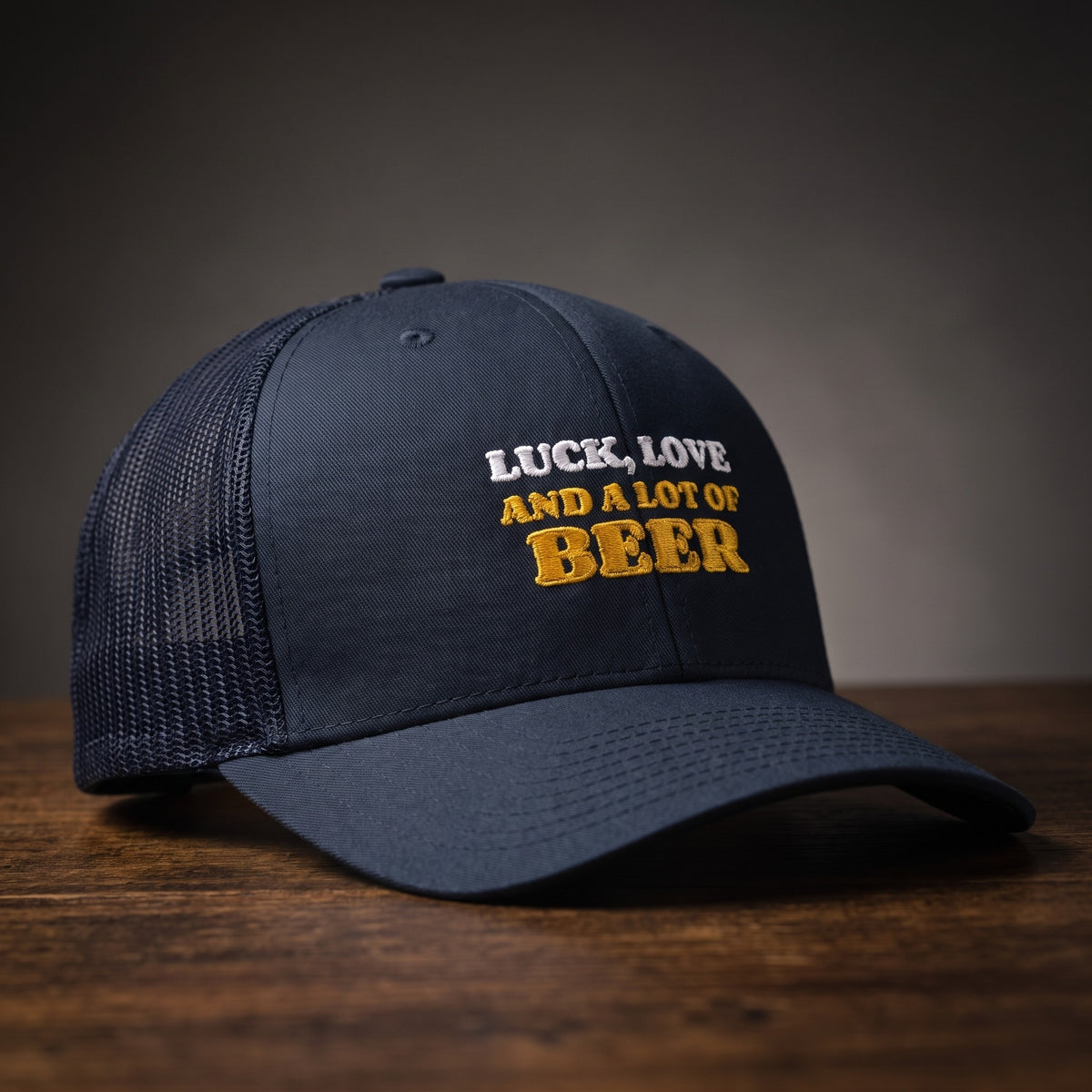 Casquette trucker bleu marine brodée « Luck, Love and a lot of Beer » en blanc et jaune doré, posée sur un bois sombre