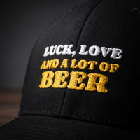 Gros plan sur la broderie « Luck, Love and a lot of Beer » en blanc et jaune doré sur casquette trucker noire