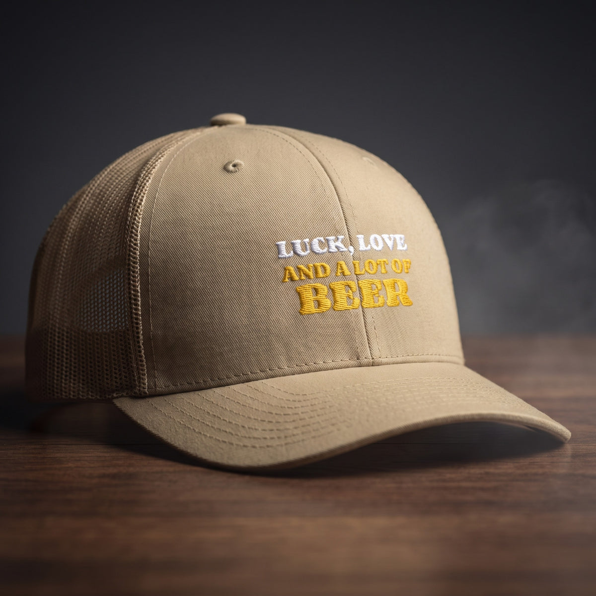 Casquette trucker kaki brodée « Luck, Love and a lot of Beer » en blanc et jaune doré, posée sur un bois sombre