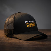Casquette trucker noire brodée « Luck, Love and a lot of Beer » en blanc et jaune doré, posée sur un bois sombre