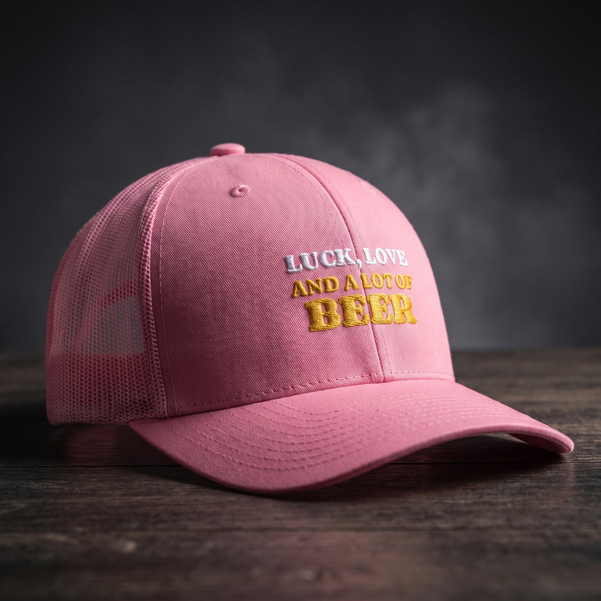 Casquette trucker rose brodée « Luck, Love and a lot of Beer » en blanc et jaune doré, posée sur un bois sombre