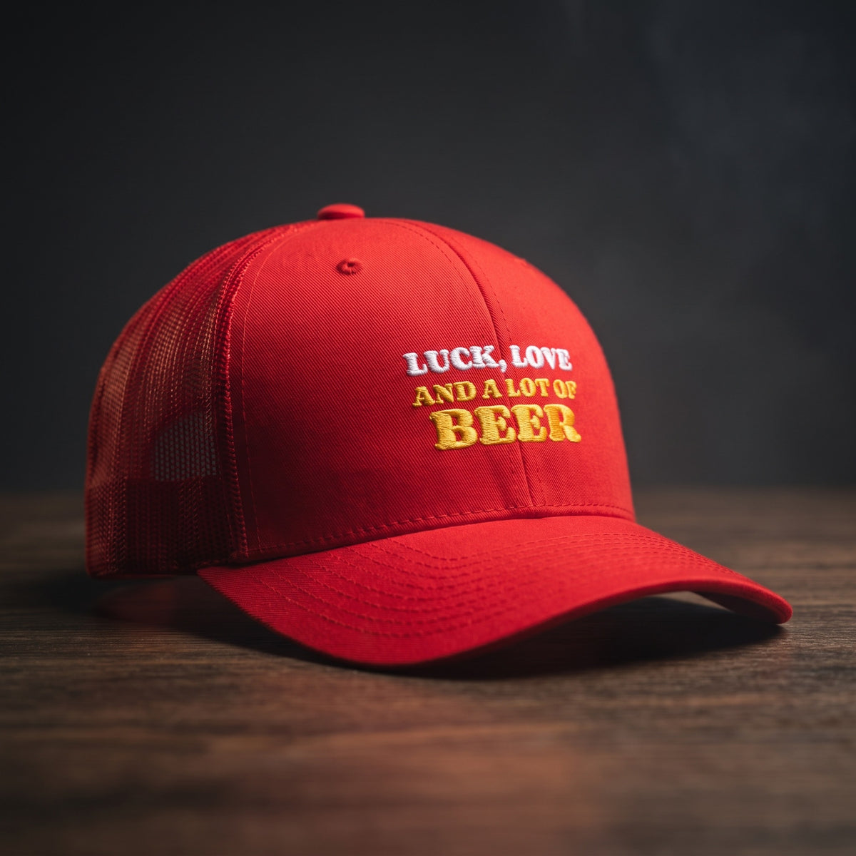 Casquette trucker rouge brodée « Luck, Love and a lot of Beer » en blanc et jaune doré, posée sur un bois sombre