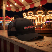Casquette trucker noire Perlinpinpin sur le comptoir d'un stand de fête foraine avec manège illuminé la nuit