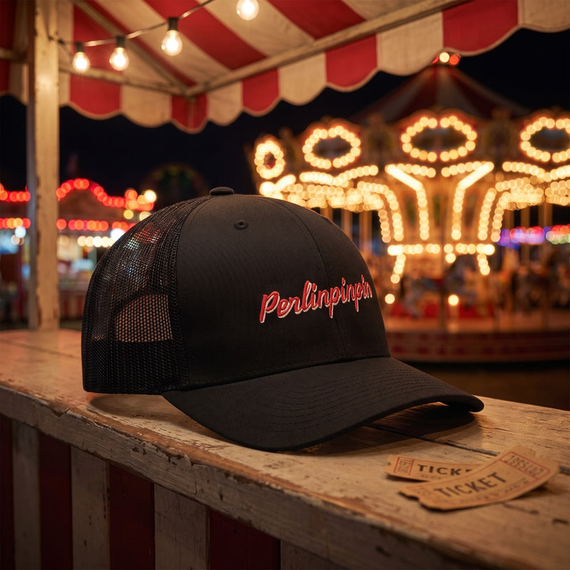 Casquette trucker noire Perlinpinpin sur le comptoir d'un stand de fête foraine avec manège illuminé la nuit