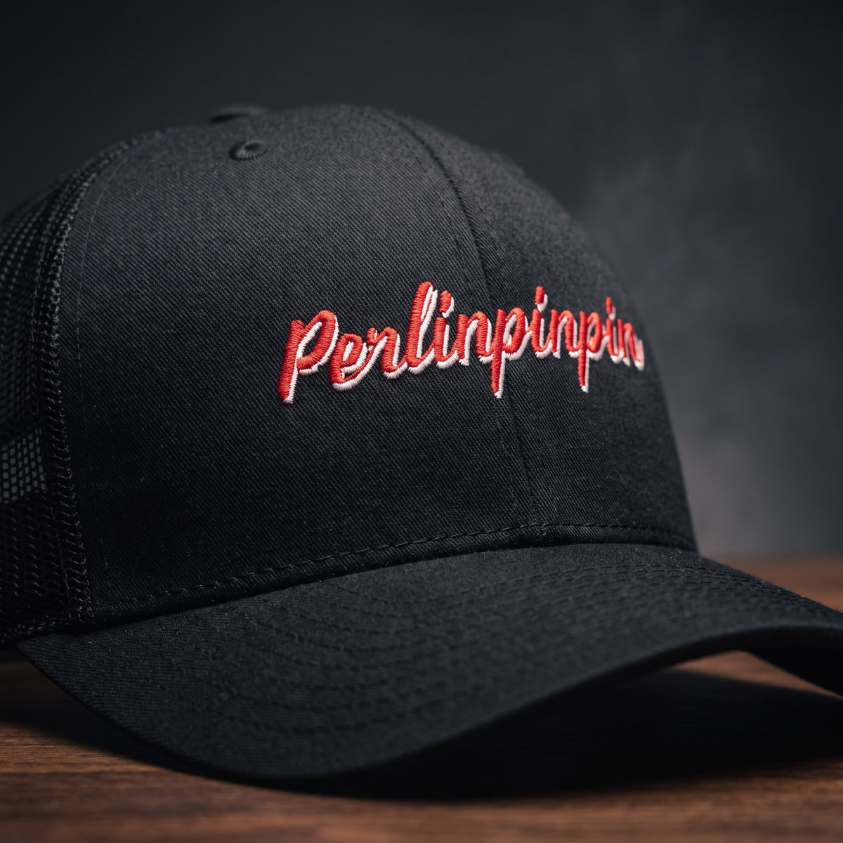 Gros plan sur la broderie Perlinpinpin en script rétro rouge et blanc sur une casquette trucker noire