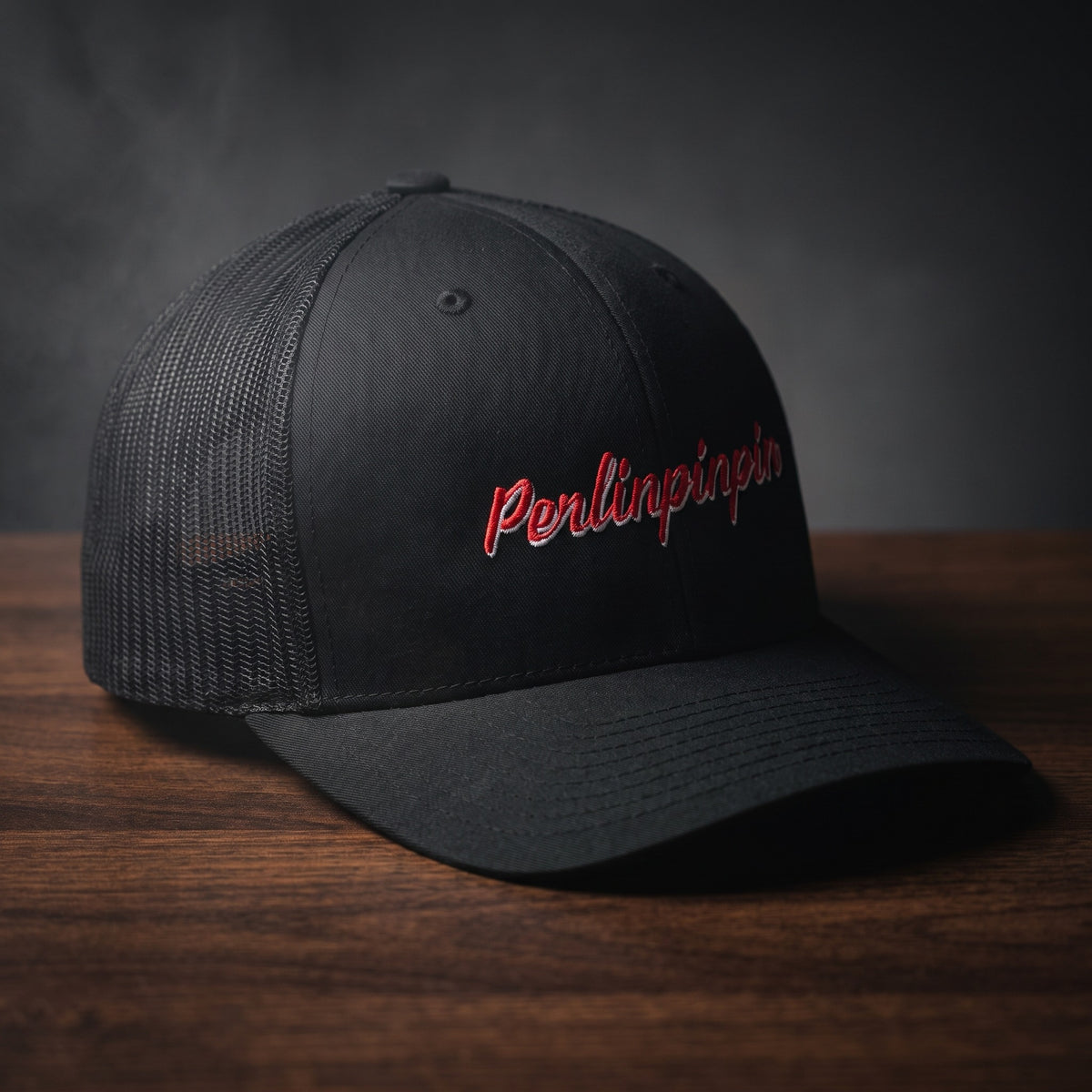 Casquette trucker noire brodée Perlinpinpin en script rétro rouge liseré blanc, posée sur une table en bois sombre