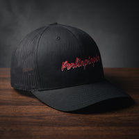 Casquette trucker noire brodée Perlinpinpin en script rétro rouge liseré blanc, posée sur une table en bois sombre