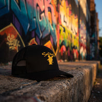 Casquette trucker Saïan Supa Crew posée sur un muret devant un mur de graffitis dans une ruelle