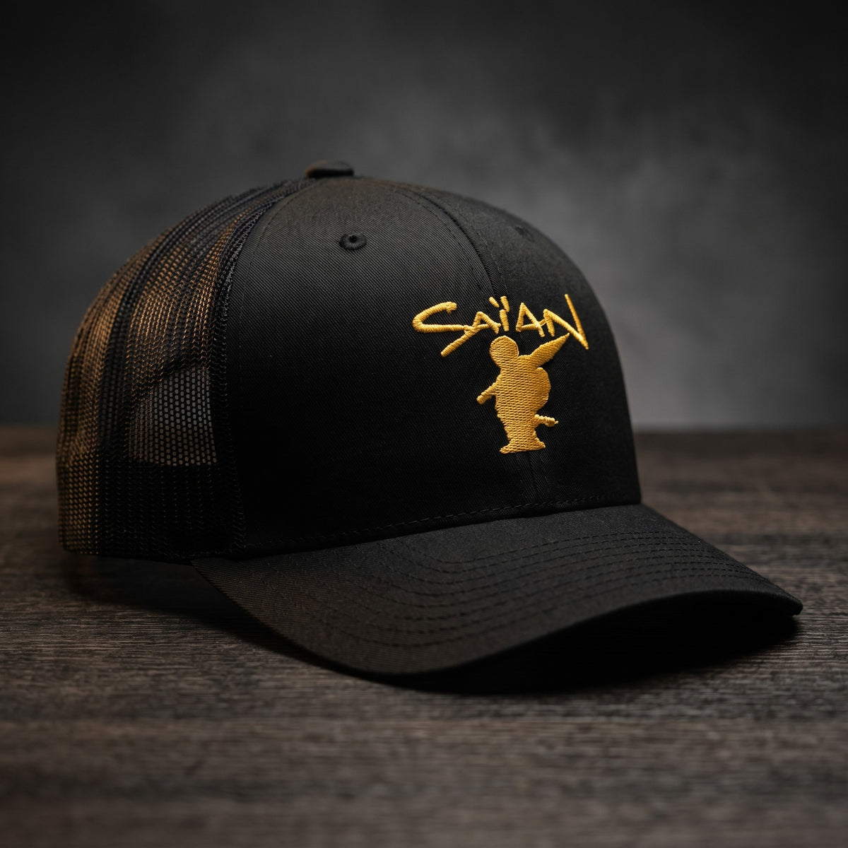 Casquette trucker noire brodée Saïan Supa Crew, lettrage jaune or, posée sur un bois sombre