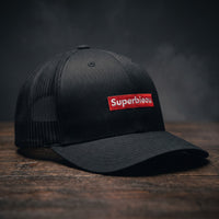Casquette trucker noire avec patch rouge brodé Superbieau, posée sur un plan en bois sombre