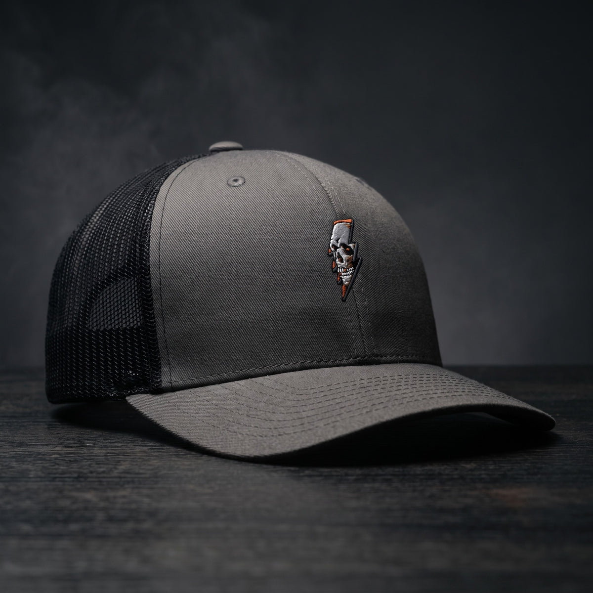 Casquette trucker Thunder Skull gris anthracite avec filet noir et crâne éclair brodé, posée sur bois sombre