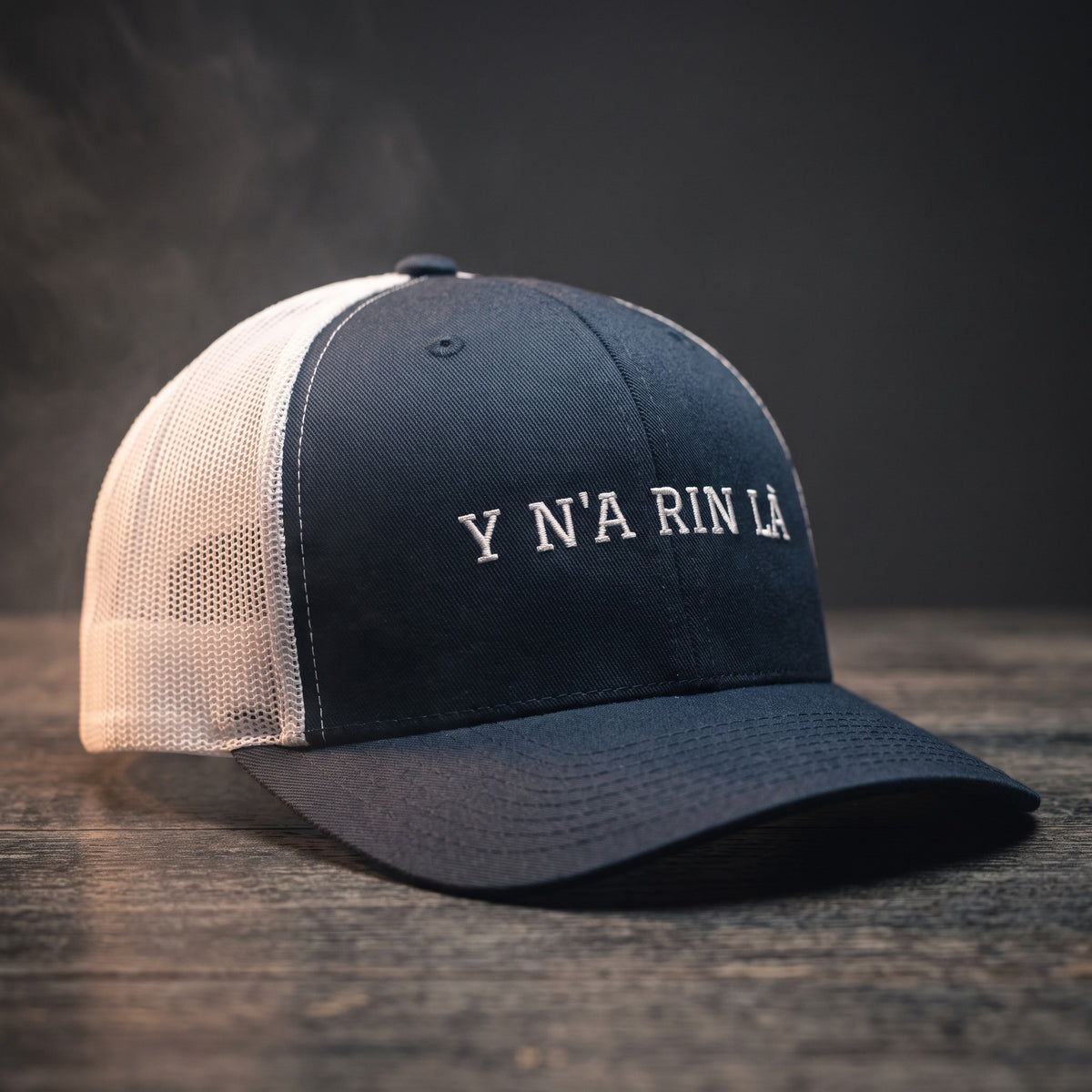 Casquette trucker bleu marine et blanche brodée « Y N'A RIN LÀ » en blanc, posée sur un bois sombre