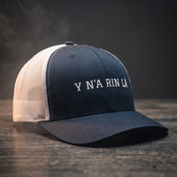 Casquette trucker bleu marine et blanche brodée « Y N'A RIN LÀ » en blanc, posée sur un bois sombre