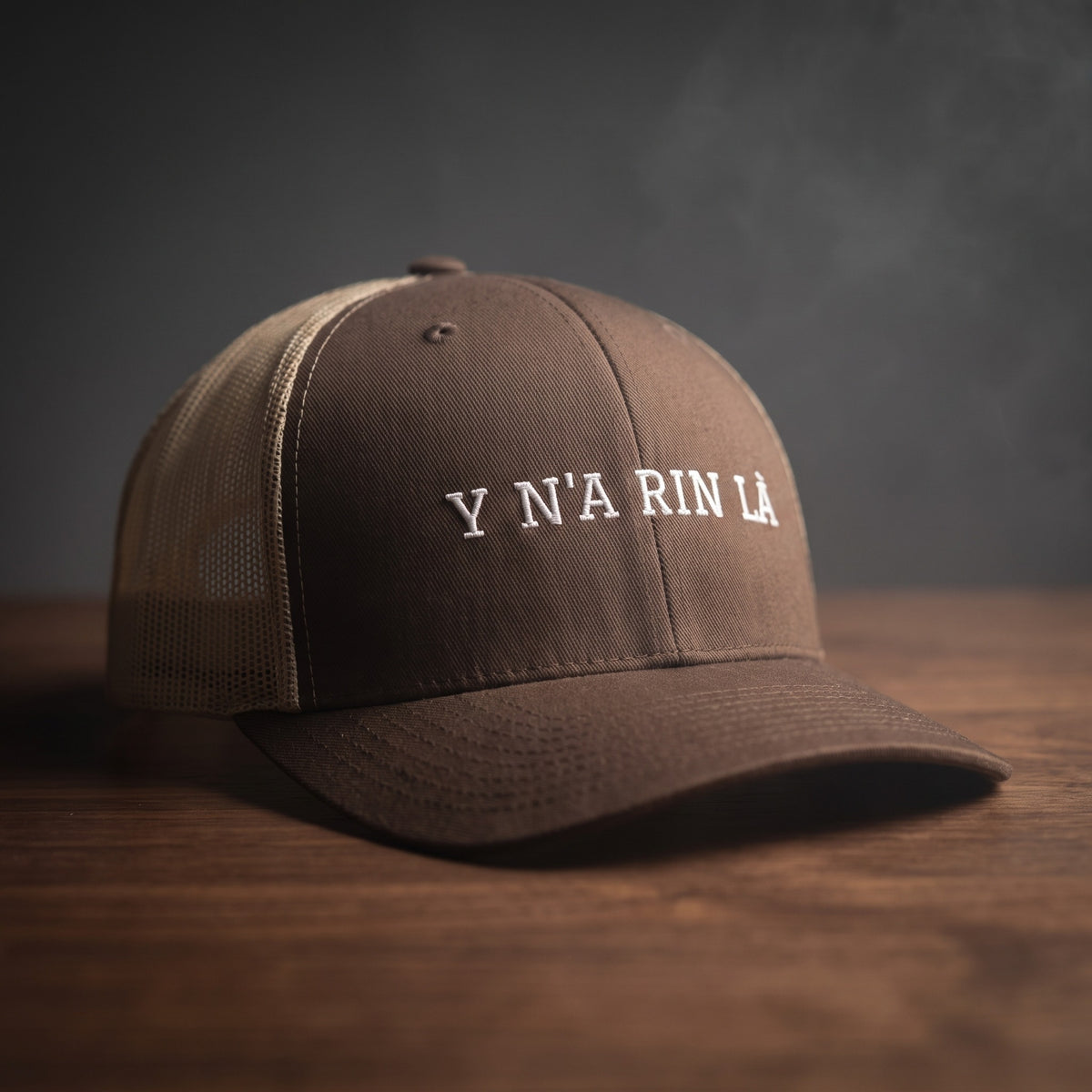 Casquette trucker marron et kaki brodée « Y N'A RIN LÀ » en blanc, posée sur un bois sombre