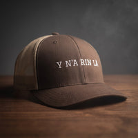 Casquette trucker marron et kaki brodée « Y N'A RIN LÀ » en blanc, posée sur un bois sombre